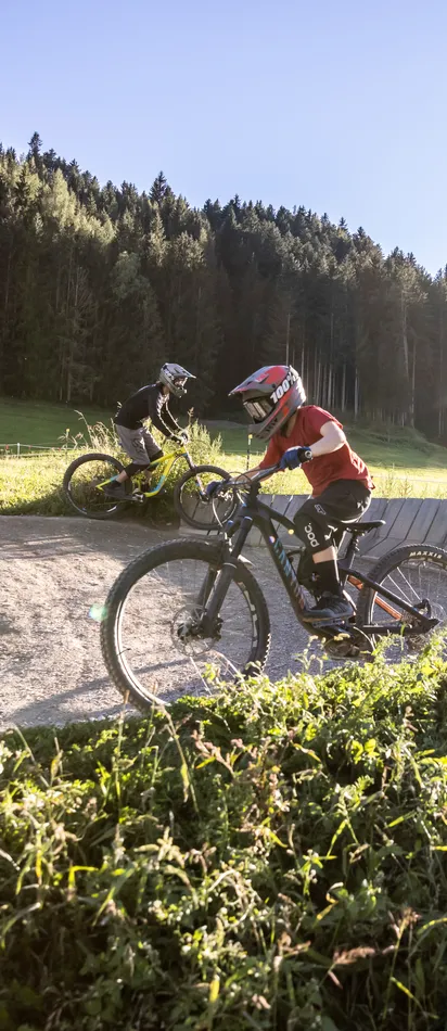 Kinder beim Mountainbiken auf einem Schotterweg mit Steilkurven in sonniger Waldlandschaft