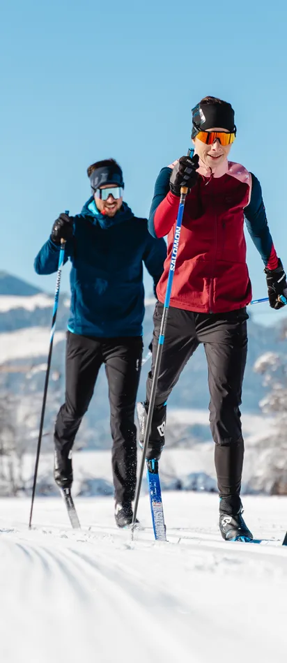 Zwei Langläufer auf einer verschneiten Loipe vor winterlicher Bergkulisse