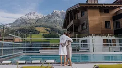 Paar in Bademänteln am Pool mit Blick auf die Alpen und ein Wellnesshotel