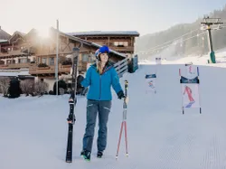 Smiling woman in ski gear holding skis and poles on a snowy slope near alpine chalets