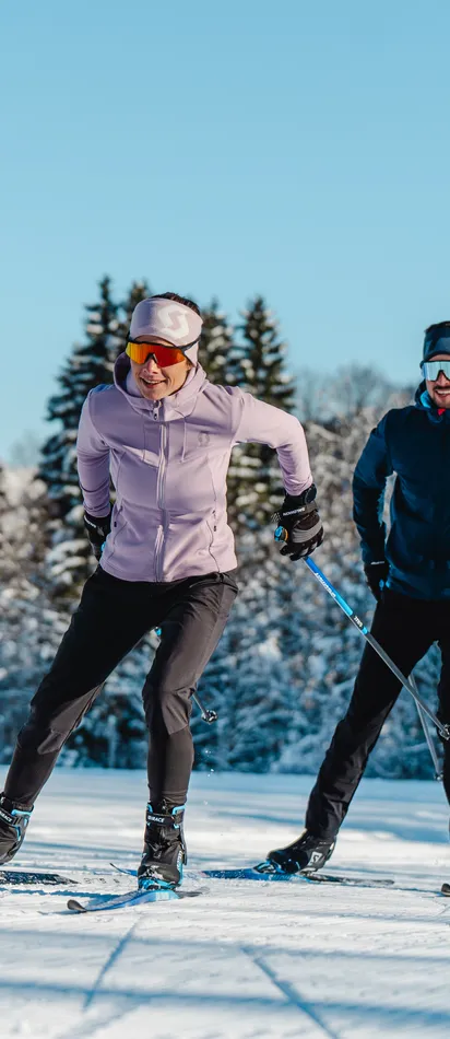 Zwei Personen beim Langlaufen auf verschneiter Loipe mit Alpen im Hintergrund