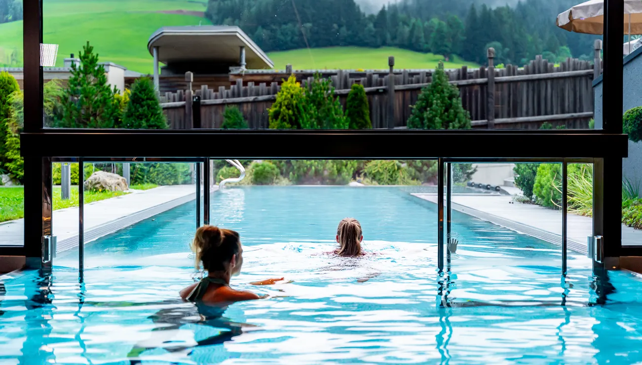 Zwei Frauen schwimmen in einem Infinity-Pool mit Blick auf grüne Hügellandschaft