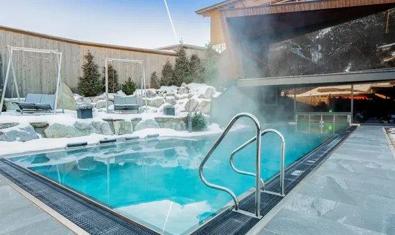Heated outdoor pool with steam rising surrounded by snow and modern alpine hotel