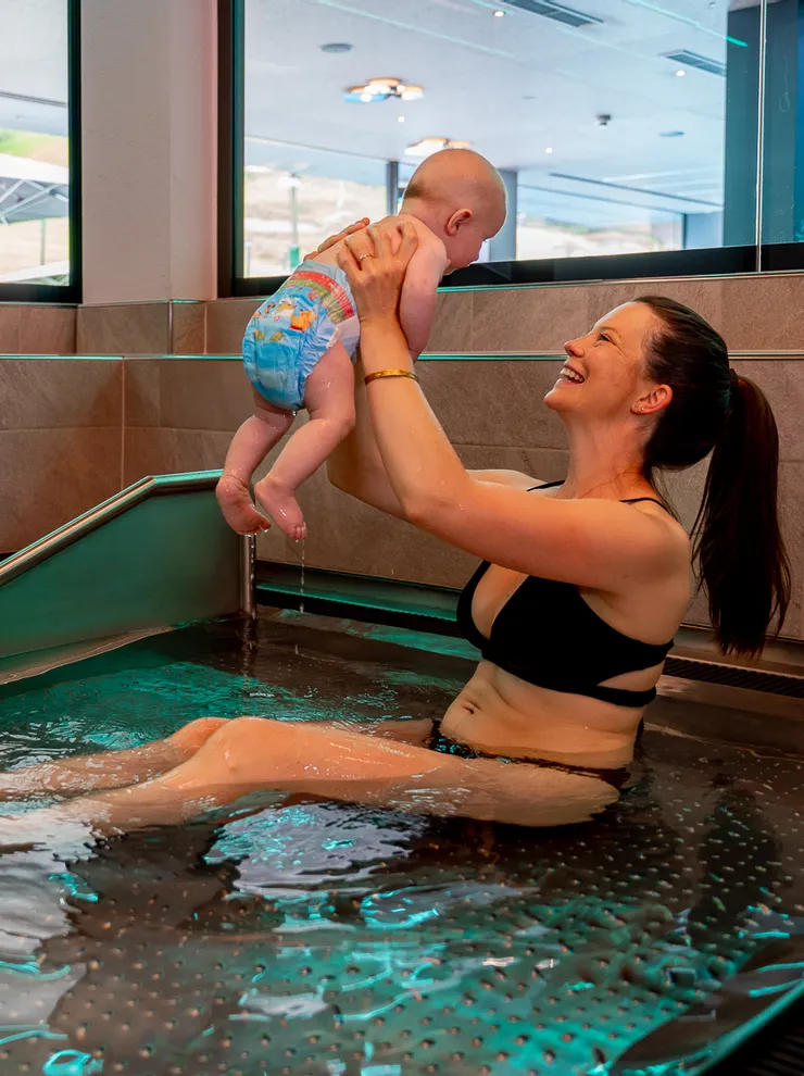 Smiling woman in bikini lifts baby in indoor pool at wellness spa