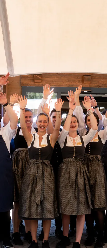 Smiling restaurant staff in traditional Bavarian attire raising hands outside a rustic venue