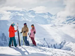 Drei Skifahrer in bunter Kleidung stehen auf verschneitem Berg mit Alpenpanorama im Hintergrund