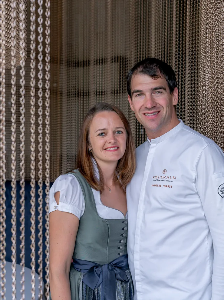 Paar vor Kettenvorhang, Mann im Kochoutfit mit Riederalm-Logo, Frau in traditionellem Dirndl