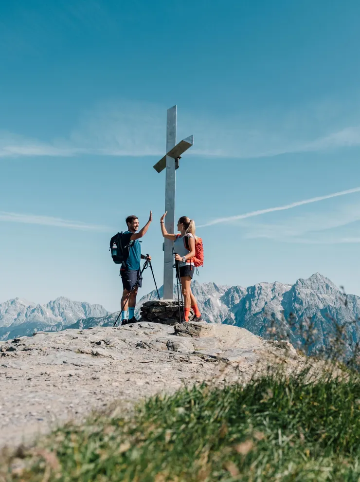 Hikers high-five at mountain summit near a cross with alpine peaks in the background