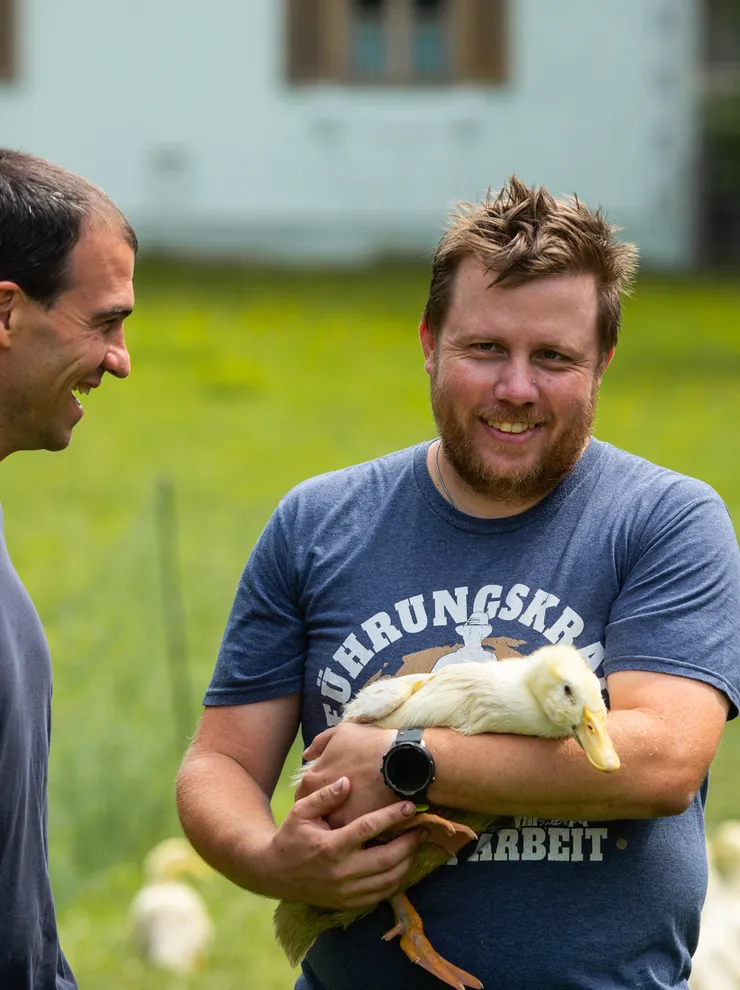 Mann hält eine Ente auf einem Bauernhof und lacht mit einem anderen Mann im Freien