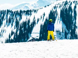 Skier in yellow pants and blue jacket overlooking snowy mountain landscape