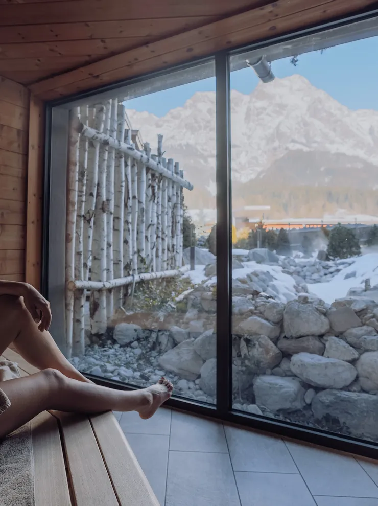 Frau in Sauna mit Blick auf verschneite Berglandschaft durch großes Fenster