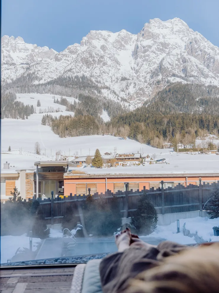 Person entspannt drinnen mit Blick auf verschneite Berge und Alpendorf durch großes Fenster