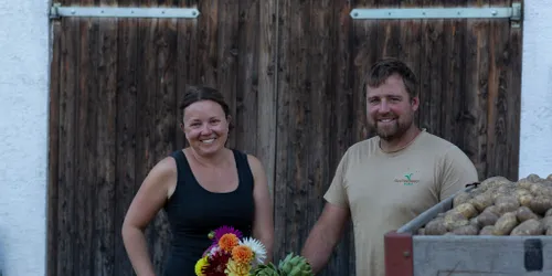 Lächelnde Landwirte neben Kisten mit Kartoffeln, Blumen und Artischocken