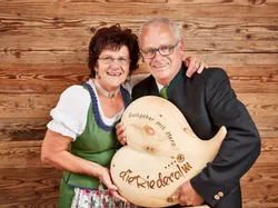 Smiling couple in traditional dress holding heart-shaped sign reading 'Gastgeber mit Herz – die Riederalm'