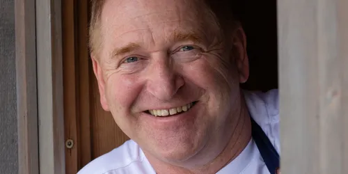 Lächelnder Koch in weißem Shirt und blauer Schürze steht in hölzernem Türrahmen