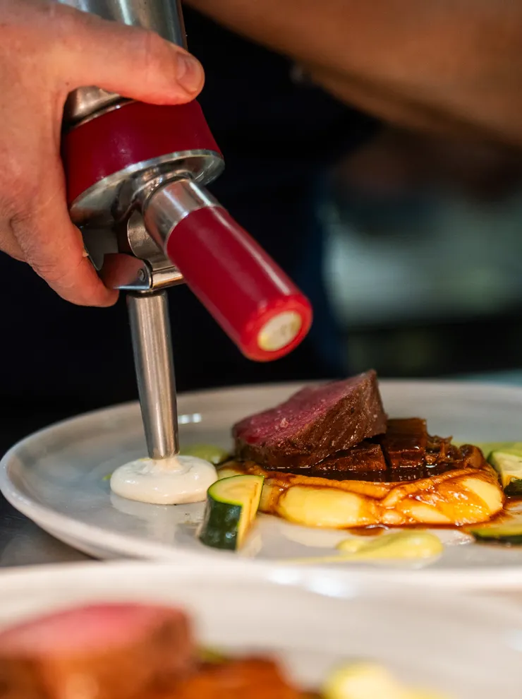 Chef plating gourmet beef dish with foam using red cream siphon in fine dining kitchen