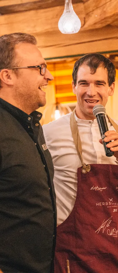 Chef with microphone smiling beside another chef at a rustic indoor culinary event