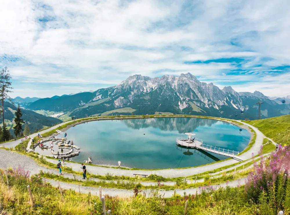 Runder Bergsee mit Steg und Aussichtspunkt vor den Alpen in der Sommerlandschaft