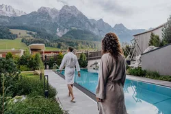 Couple in robes walking by outdoor pool at alpine wellness resort with mountain view