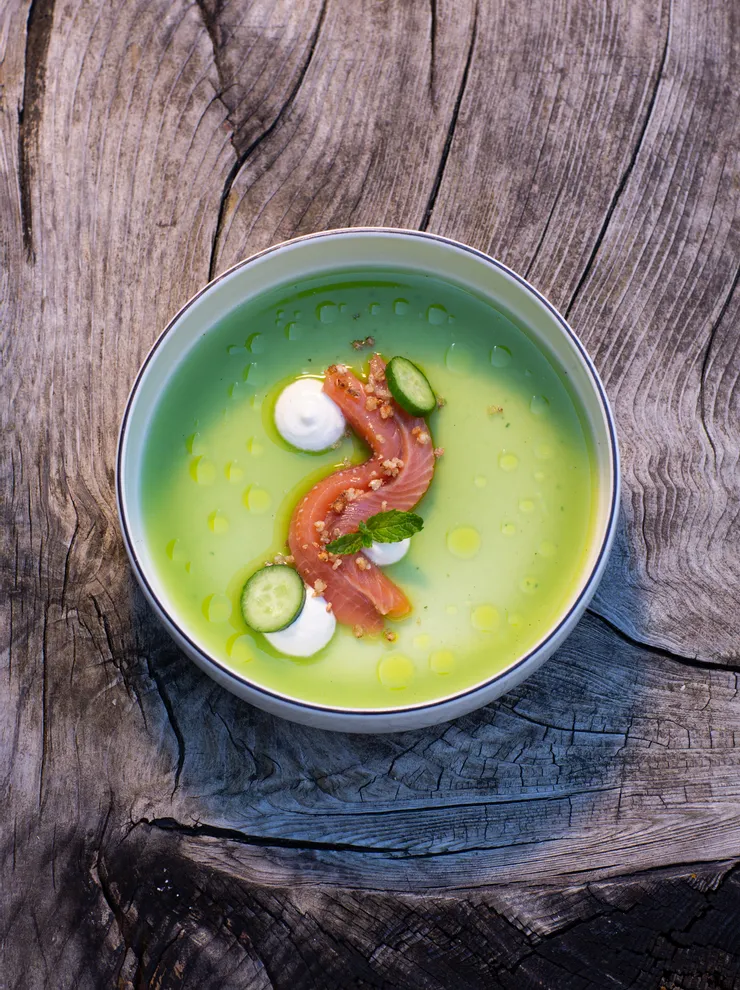 Green cucumber soup with raw salmon, cream, and herbs served in a bowl on rustic wood