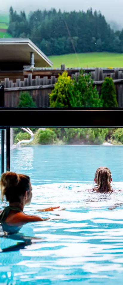 Zwei Frauen schwimmen in einem Infinity-Pool mit Blick auf grüne Hügellandschaft