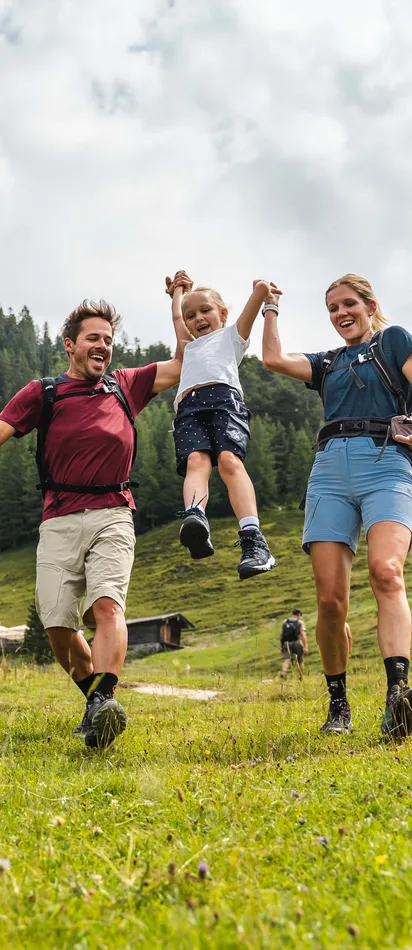 Familie wandert durch grüne Alpenwiese mit Kind in der Luft und Bergen im Hintergrund