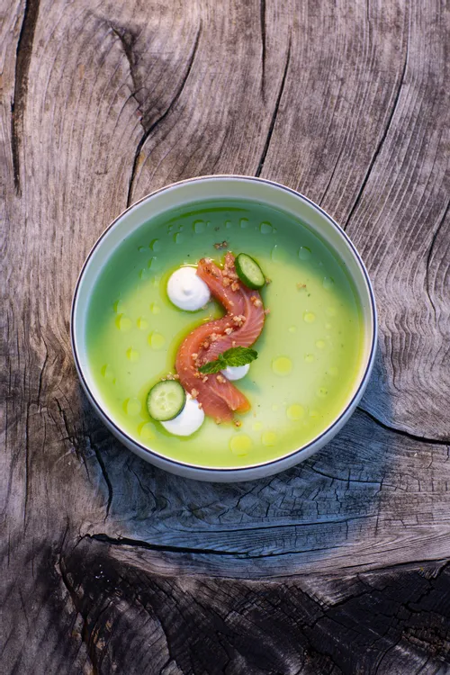Green cucumber soup with raw salmon, cream, and herbs served in a bowl on rustic wood