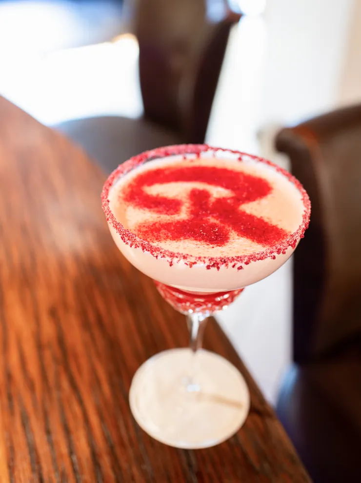 Rot-weißer Cocktail im Coupette-Glas mit Zuckerrand und Buchstabe R obendrauf