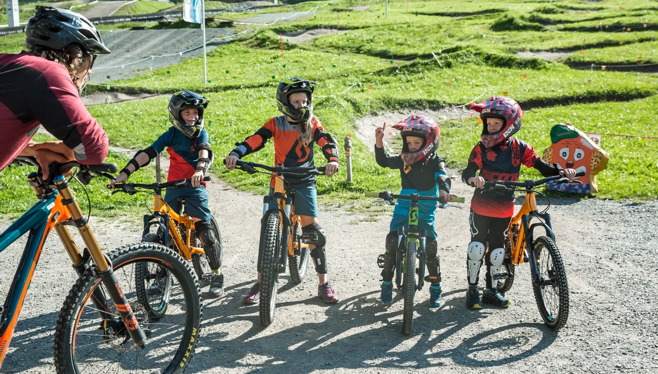 Kinder in Schutzausrüstung auf Mountainbikes sprechen mit Erwachsenen im Bikepark