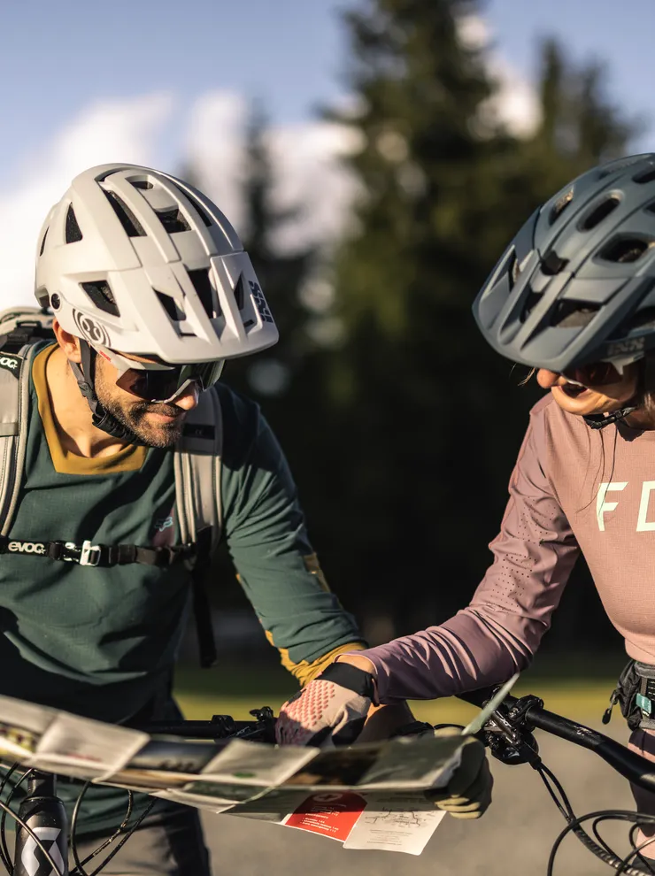 Zwei Mountainbiker mit Helmen lesen eine Karte auf sonnigem Weg vor Alpenkulisse