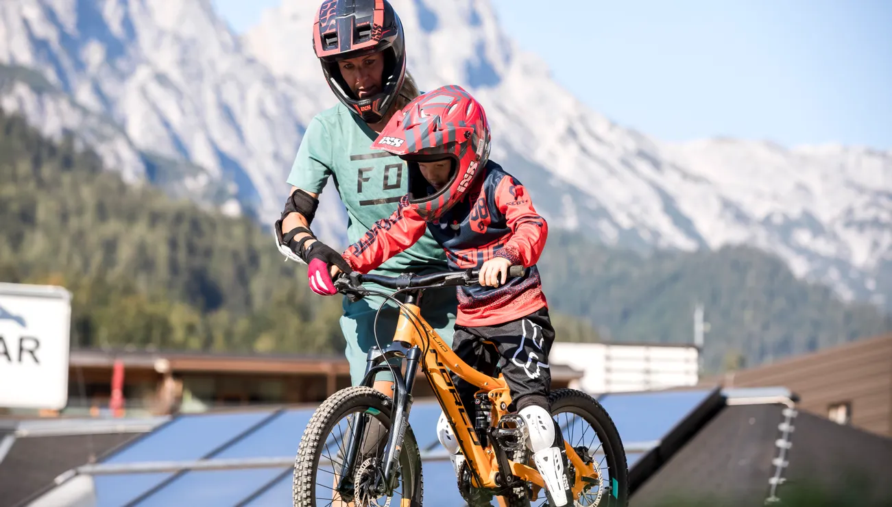 Erwachsener bringt Kind Mountainbiken auf einem Trail mit Alpenpanorama bei