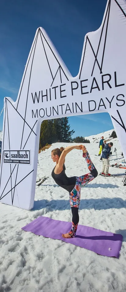 Frau macht Yoga im Schnee bei den White Pearl Mountain Days in Saalbach mit Skifahrern im Hintergrund