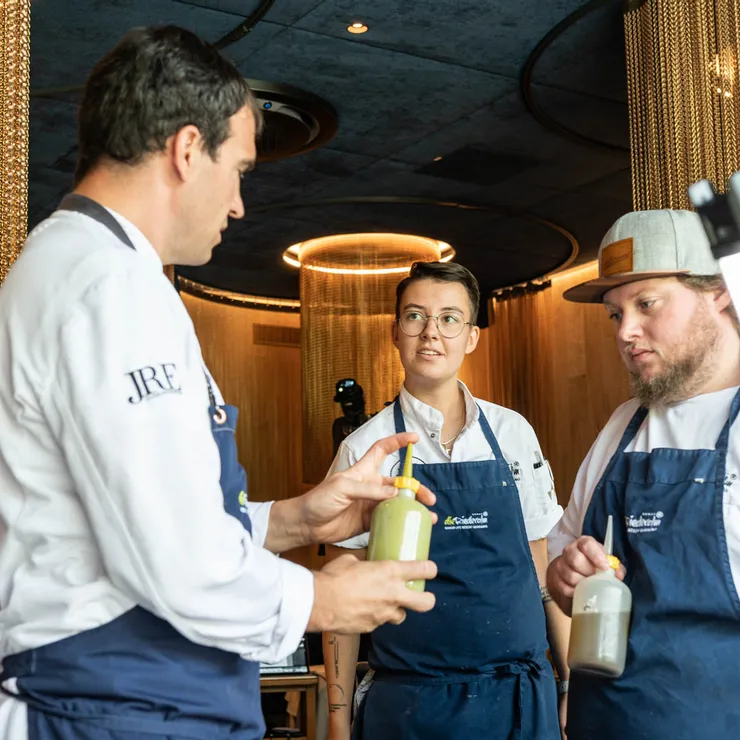Three chefs discussing sauce preparation in a modern restaurant kitchen