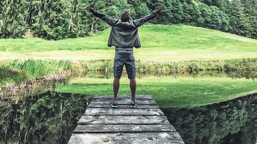 Man standing on wooden dock with arms raised overlooking forest and grassy field