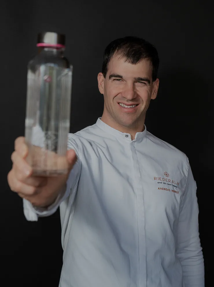 Lächelnder Koch in weißer Uniform hält eine klare Wasserflasche vor dunklem Hintergrund