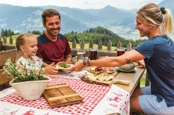 Familie genießt traditionelles Alpen-Mittagessen auf Holzterrasse mit Bergblick