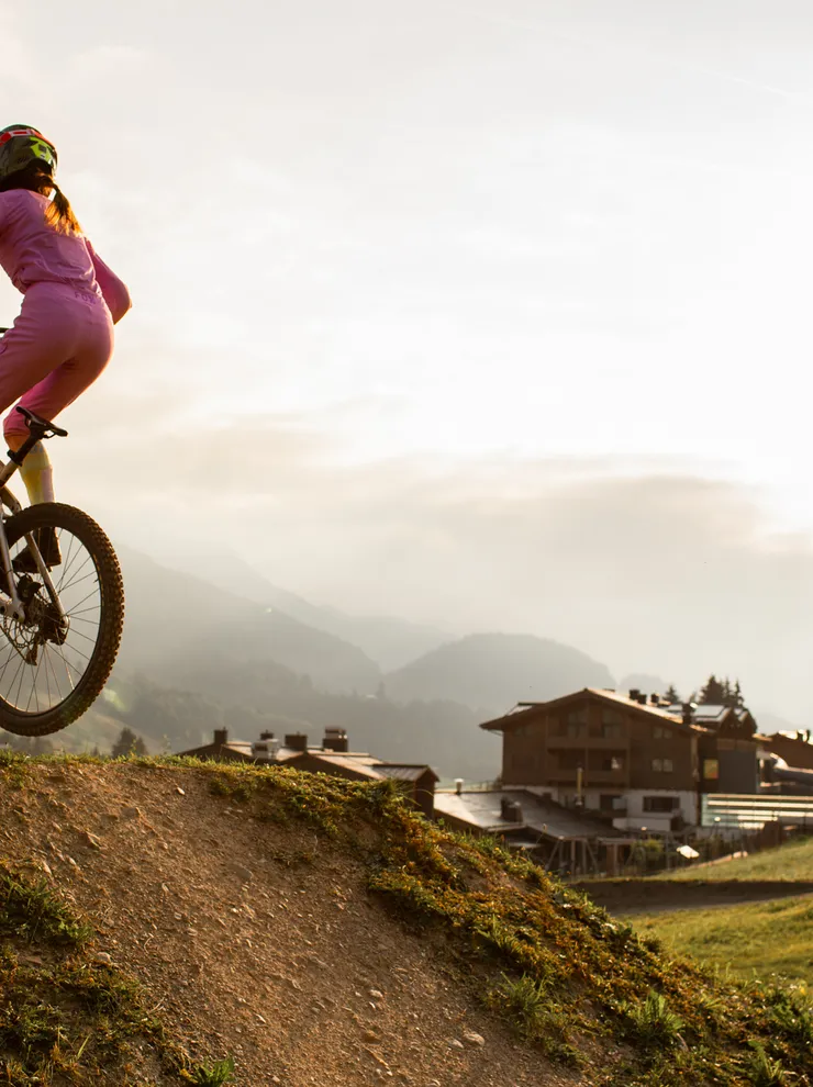 Frau in pinkem Outfit springt mit Mountainbike von Erdhügel im alpinen Sonnenaufgang