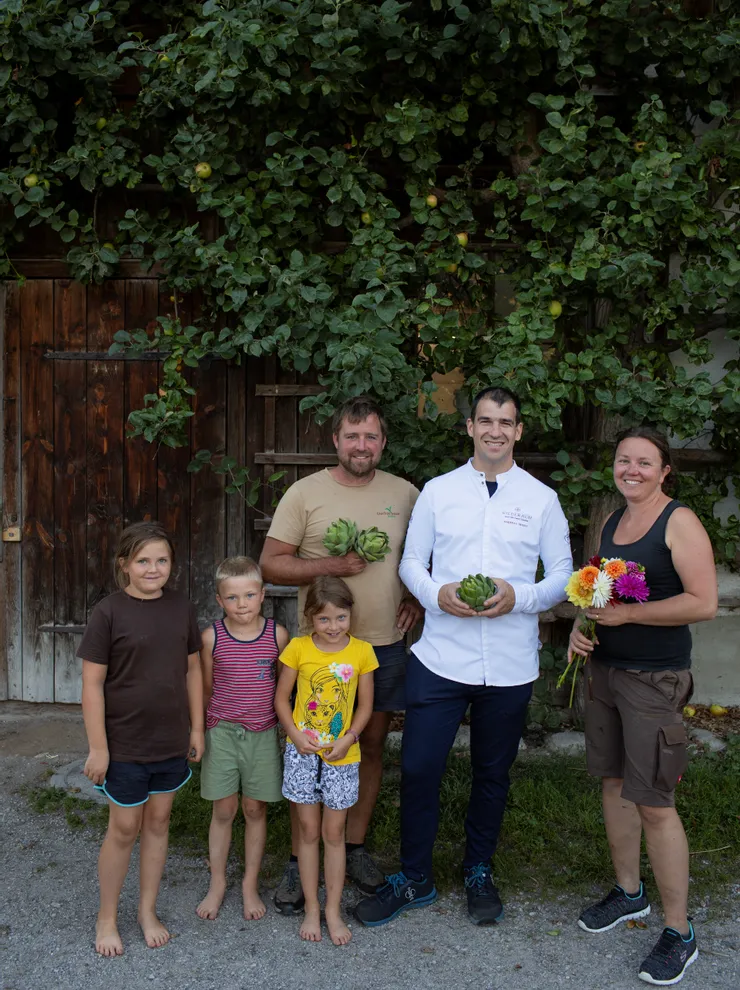 Familie mit Koch hält Artischocken und Blumen vor rustikalem Holzhaus mit grünem Laub