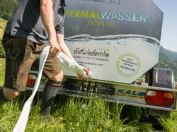 Man in traditional attire connects hose to thermal water tank trailer in green meadow