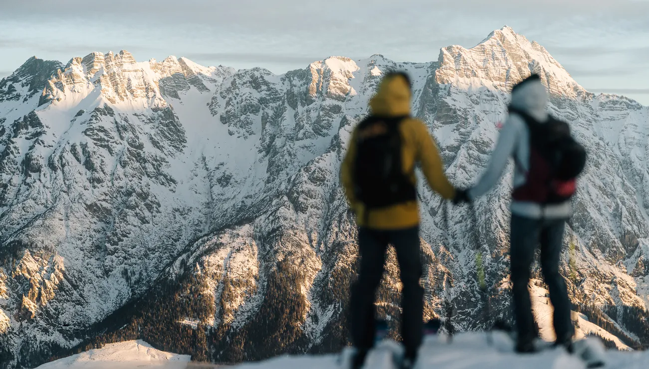 Zwei Wanderer mit Winterkleidung blicken auf verschneite Alpenberge im Morgenlicht