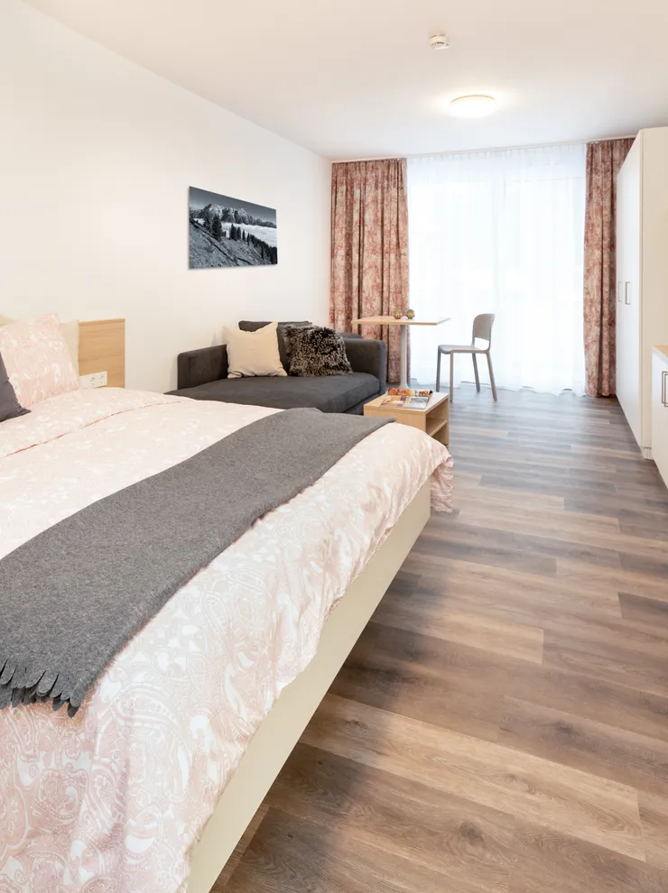 Modern hotel room with double bed, sofa, TV and kitchenette on wooden floor