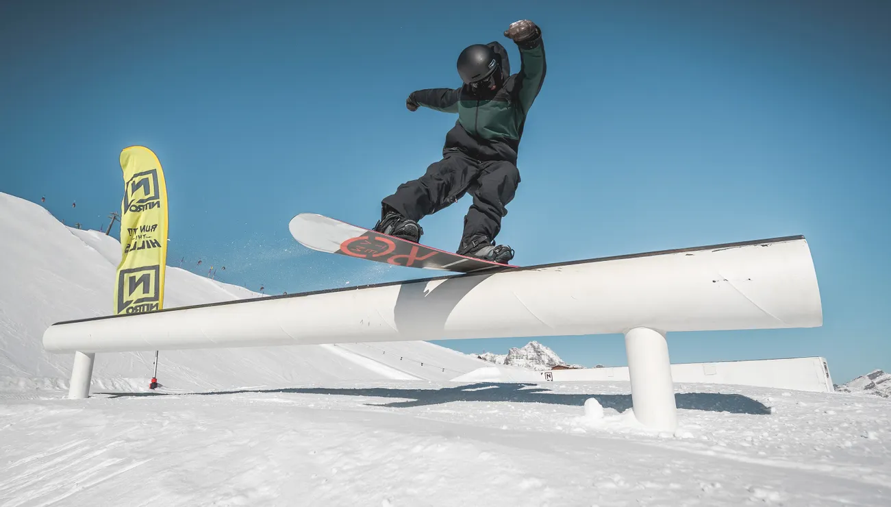 Snowboarder performing a rail slide trick in a sunny terrain park