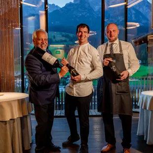 Three men in fine dining restaurant holding wine bottles with mountain view in background