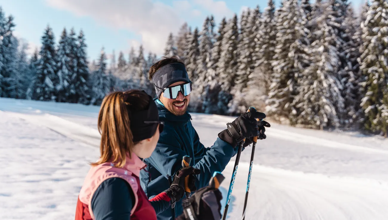 Zwei Langläufer unterhalten sich auf einer verschneiten Waldloipe bei Sonnenlicht