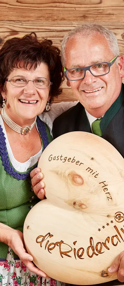 Lächelndes Paar in Tracht hält Herzschild mit Schriftzug 'Gastgeber mit Herz – die Riederalm'
