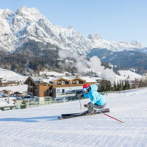 Skier in blue jacket carving on groomed slope with snowy Alps and chalet in background