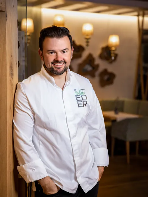 Mann im weißen Kochhemd lehnt lächelnd an einer Holzwand im Restaurant