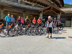 Group of cyclists with mountain bikes gathered outside a rustic lodge in sunny weather