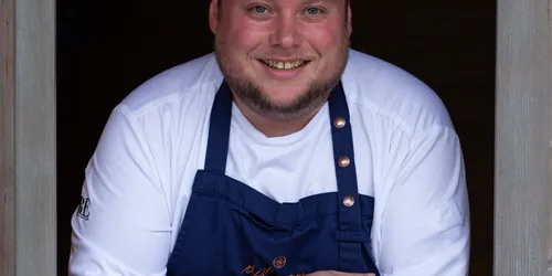 Lächelnder Koch mit blauer Schürze und Mütze lehnt im hölzernen Fensterrahmen