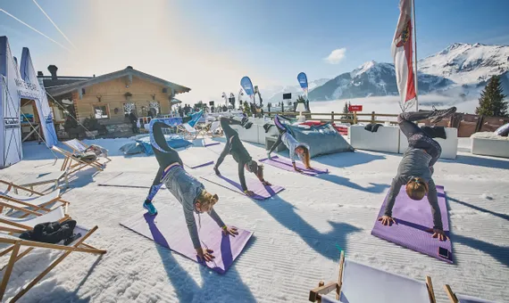 Frauen machen Outdoor-Yoga im Schnee in den Alpen bei einer Skihütte unter blauem Himmel